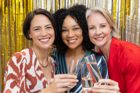 Diverse women friends raising champagne flutes smiling posing at party in party wear foil fringe