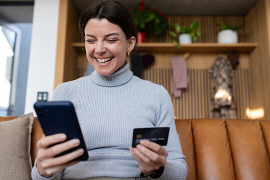 Woman sitting on caramel-brown leather sofa at home using smartphone and holding black payment card