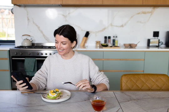Woman sitting at marble island holding smartphone and fork eating avocado toast with poached egg