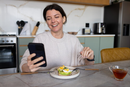 Woman in 30s in tan sweater sitting kitchen holding smartphone, tea and egg-topped avocado toast
