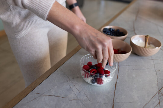 Adult female placing raspberry into glass bowl of yogurt and berries on gray stone countertop