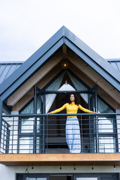 African woman standing on gabled balcony, wearing yellow top, blue jeans, resting on metal railing