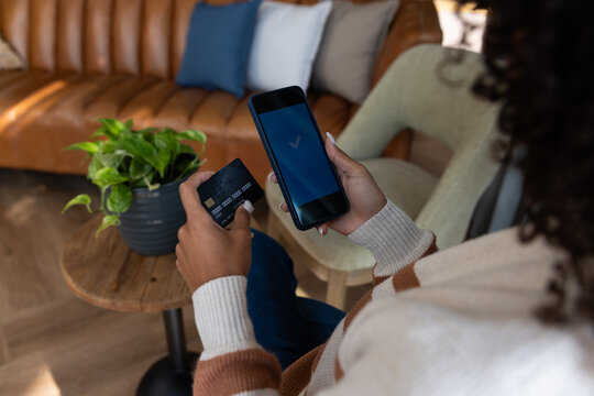 Woman 20s sitting at home holding smartphone with checkmark and black card, wearing striped sweater