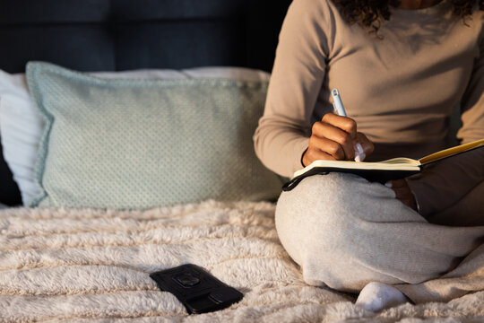 Adult African American woman in beige top gray sweatpants writing in notebook on bed holding pen