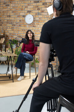 Female guest sitting and male operator filming camera in studio, guest facing microphone