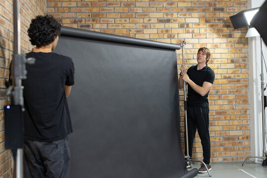 Two men installing large black paper backdrop in studio, holding support pole and softbox