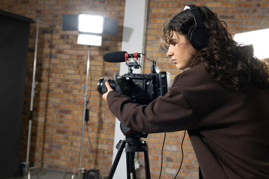 Woman operating camera with shotgun microphone on tripod in studio, wearing brown hoodie
