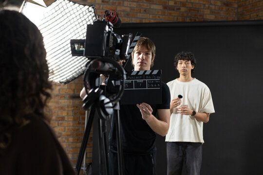 Diverse crew holding clapperboard and operating camera on tripod in studio with softbox light