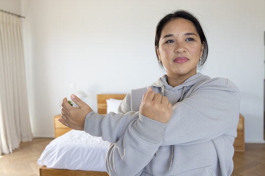 Female wearing grey hoodie stretching arms across chest near wooden bedframe and lamp in bedroom