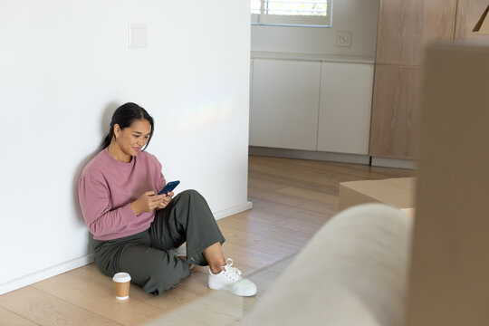Asian woman sitting on floor holding smartphone, wearing mauve top, coffee cup, boxes, copy space