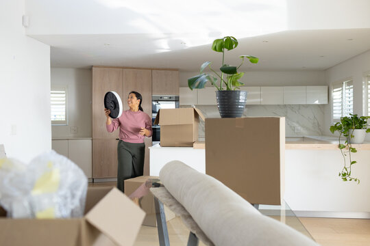 Asian woman standing holding black clock in pink top, olive pants amid boxes in open-plan kitchen