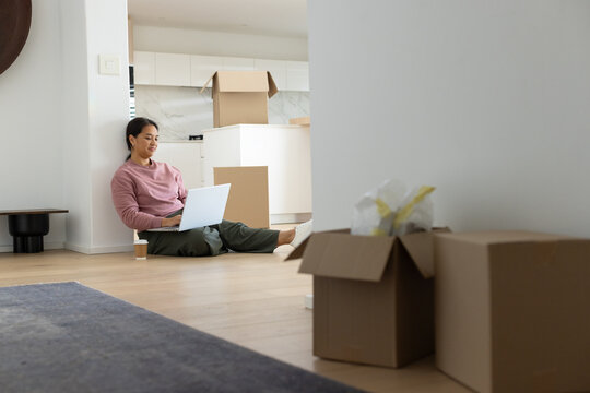 Asian woman sitting at home among moving boxes using laptop with coffee, copy space