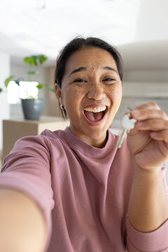 Asian woman taking front-facing selfie holding keys up in kitchen with boxes wearing mauve top