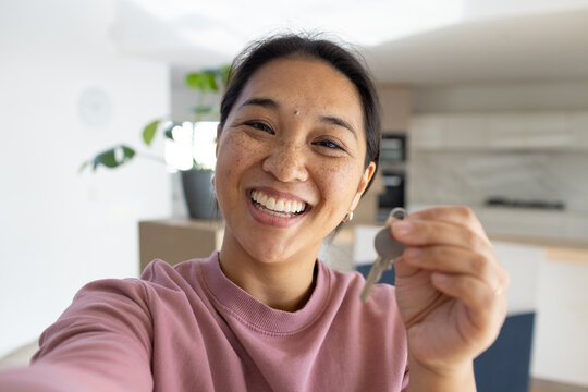 Asian woman smiling while holding metal key on keyring in home kitchen wearing pink crewneck
