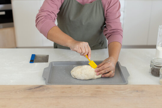 Asian woman wearing olive green apron shaping loaf dough on gray baking tray at kitchen counter