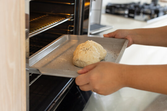 Woman placing metal baking tray with seeded dough into oven in home kitchen near gas stovetop