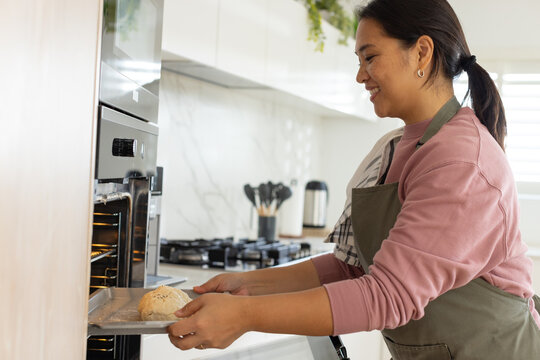 Asian woman placing round dough on baking tray into built-in oven in home kitchen, copy space