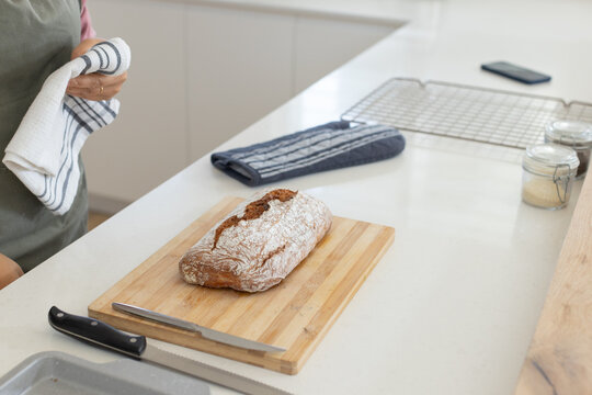 Woman wearing green apron holding white striped towel while slicing rustic bread at kitchen counter