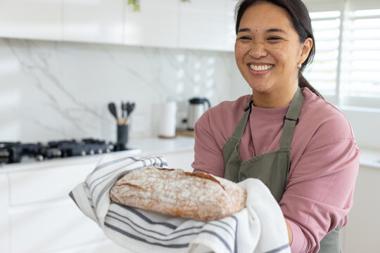 Asian woman smiling holding baked loaf wrapped in striped towel, wearing green apron in kitchen