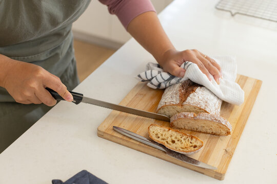 Asian woman wearing green apron slicing rustic floured loaf on white countertop with serrated knife