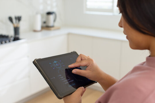 Adult Asian woman standing in home kitchen holding tablet in case tracing schematic, copy space