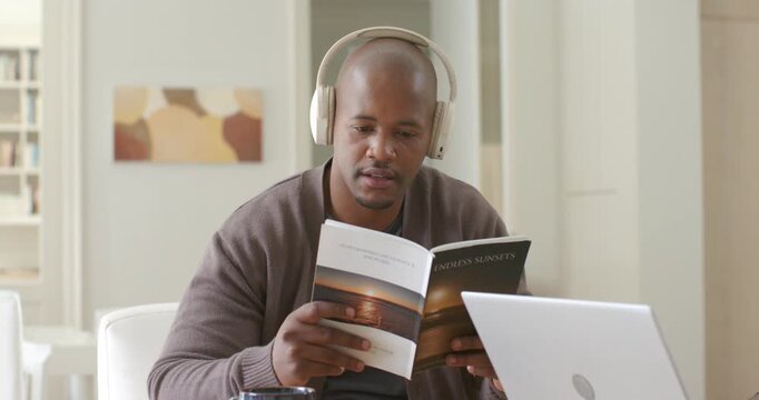African man at table opening ENDLESS SUNSETS booklet following audio on headphones glancing laptop