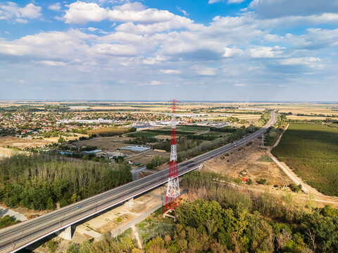 Aerial view of the M8 Pentele Bridge approach with traffic and high-voltage pylons near Dunavecse, Hungary, bordered by farmland, trees and a low-rise town.