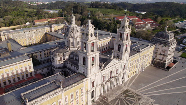 Aerial view of the historic Mafra National Palace with its grand basilica, twin bell towers, and expansive courtyard, surrounded by lush green hills in Mafra, Lisbon, Portugal.