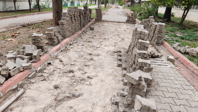 An outdoor walkway undergoing renovation, with interlocking paving stones stacked in piles along the borders, exposing the sandy ground underneath.