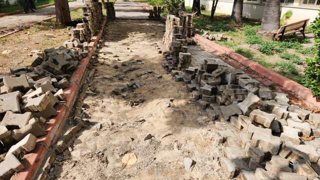 An outdoor walkway undergoing renovation, with interlocking paving stones stacked in piles along the borders, exposing the sandy ground underneath.