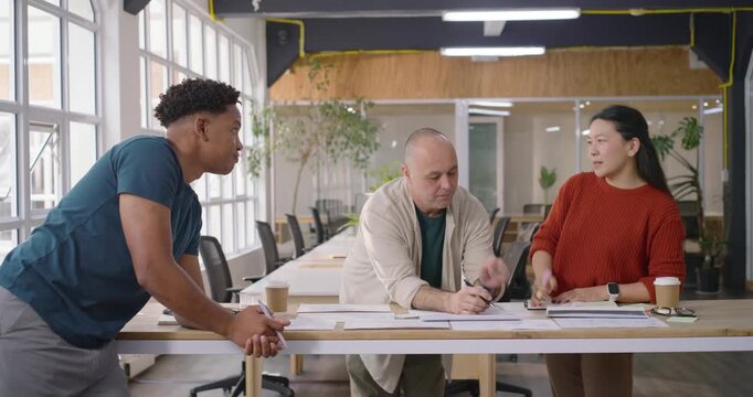 Team standing at long table in office inspecting layouts, pointing pen, noting to finalize designs