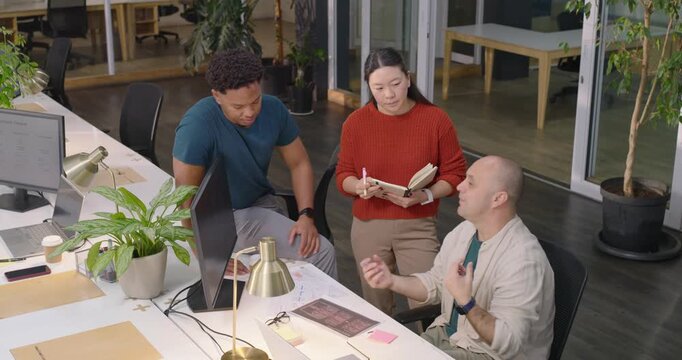Diverse coworkers reviewing layouts at desk after seated lead pointing, noting feedback for project