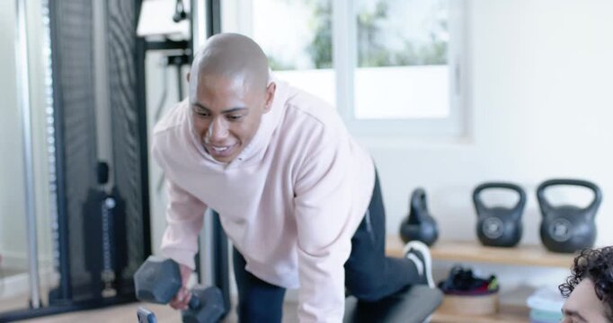 Male friends showing phone, exerciser performing one-arm dumbbell rows on bench following coach