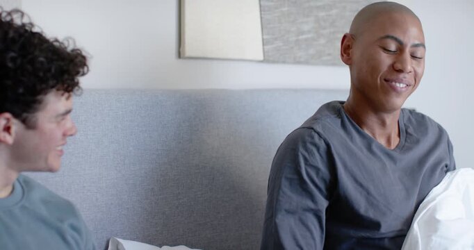 Mid 20s men sitting on bed, left man holding remote initiating right man waking, stretching