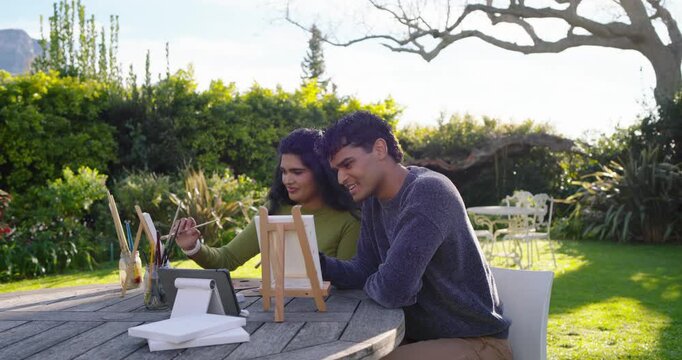 Couple painting together at wooden table in garden, using tablet reference and sharing brushes