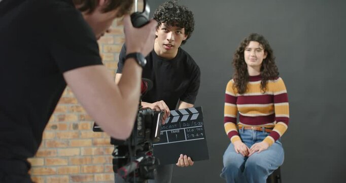 Film crew recording set take, clapper snapping slate, camera framing woman striped sweater on stool