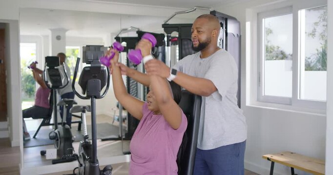 African American woman pressing purple weights on bench at home while trainer spotting