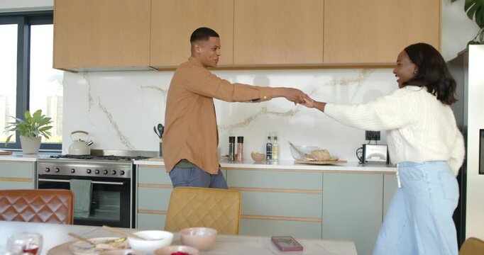 African American couple taking hands, twirling, dancing affectionately in kitchen near smartphone