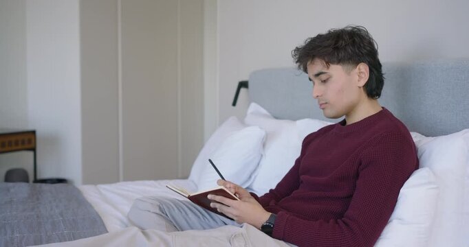 African American man journaling in brown notebook on bed after reading notes with pencil