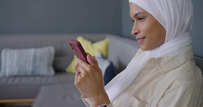 African American woman in white hijab holding phone at chest, checking messages on sofa, copy space
