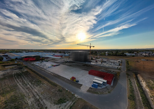 Aerial view of a warehouse construction site with a tower crane, steel framework, materials and access roads in Dunaharaszti, Hungary, in an industrial zone at sunset.
