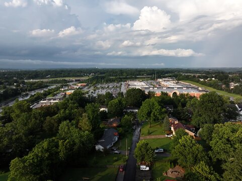 Aerial view of a shopping center and parking lot surrounded by lush green trees and residential houses under a dramatic cloudy sky in Crestview Hills, Kentucky, United States.