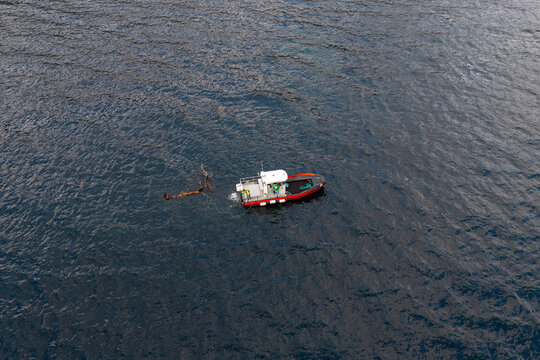 Aerial view of a red workboat towing a large tree away from a fish farm in the deep blue waters of a fjord in Fjord, More og Romsdal, Norway.