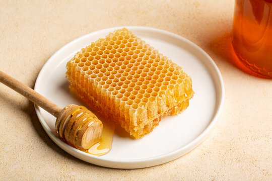 Honeycomb filled with golden raw honey served on a white plate with a wooden honey dipper and honey jar in the background. Rustic natural food still life photographed from a 45-degree angle.