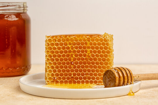 Natural honeycomb standing vertically on a white ceramic plate, filled with golden raw honey, accompanied by a wooden honey dipper and honey packaging. Rustic still life