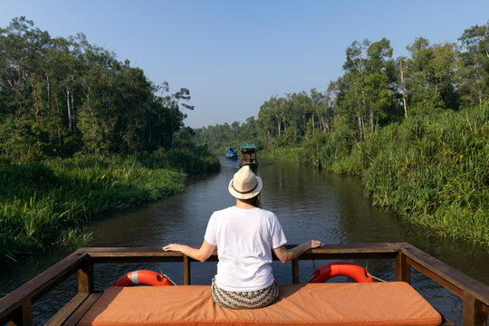 An Indonesian *klotok* boat is navigating a jungle river. A female tourist is seated on the boat, facing the stern. Impenetrable groves of trees and lush green grass line the banks. Blue sky, clouds.