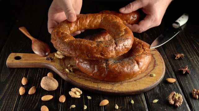 Hands lift one ring shaped loaf from stacked golden baked breads on a wooden board surrounded by nuts and spices, overhead food preparation scene