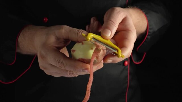Hands peeling a potato with a yellow vegetable peeler, removing a long strip of skin against a dark background during food preparation