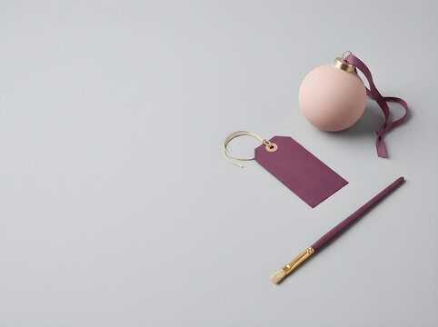 Minimalist holiday still life with pink Christmas bauble and purple gift tag, elegant festive craft scene with paintbrush, copy space on grey