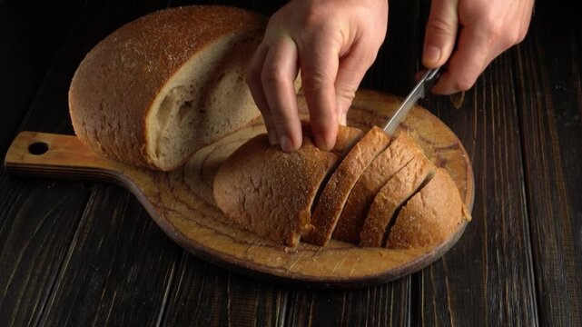 Hands use a sharp bladed knife to slice crusty bread into even pieces on a round wooden board, while another loaf lies behind on a dark, rustic style table